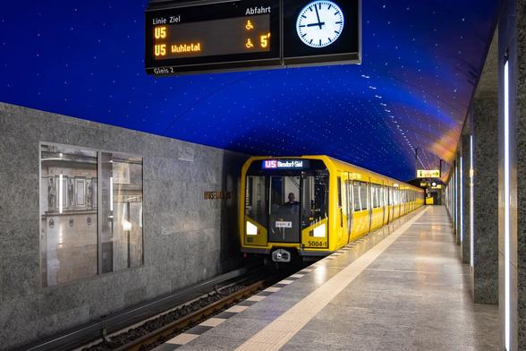 Wide-angle view of one of the two platforms at Museumsinsel U-Bahn Station in Berlin. Some pictures of the Crown Prince's Palace are decorating the wall made of grey stone. A yellow metro train is entering the station under a painted dark blue starry ceiling with a curvy shape.