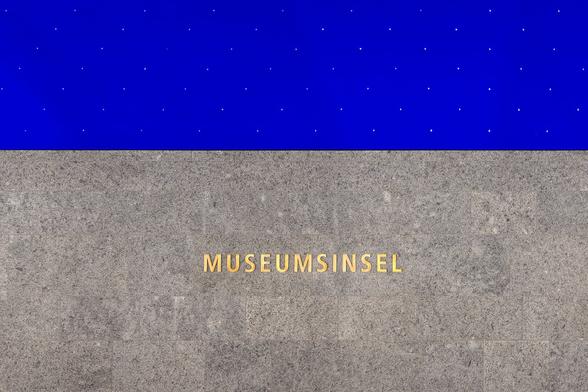 Wall at Museumsinsel U-Bahn Station in Berlin featuring the station name in golden letters over grey stones and a dark blue starry ceiling.