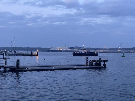 Kieler Förde am Abend. Im Vordergrund ein Rudersteg, dahinter die Schwentinefähre und dahinter am Ostufer die Litauenfähre. Der Himmel ist grau, das Wasser hat Miniaturwellenberge und die Lichter der Laternen auf dem am linken Rande sichtbaren Reventlouanleger sind gerade angegangen.
