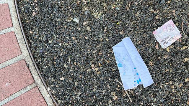 a faded label for generic non-dairy creamer sits on asphalt by a folded piece of note paper with illegible blue ink soaked through the underside. a curved portion of a brick sidewalk can be seen opposite of the label.