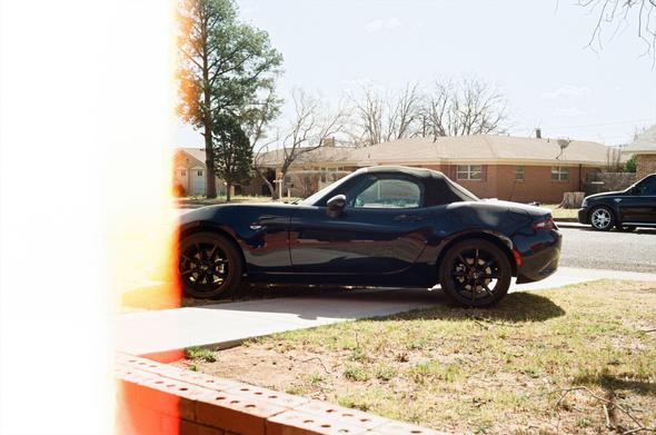 Dark blue Mazda MX-5 parked on a front lawn. Photo was taken using the beginning edge of a roll of film, so it has a color cast and white edge from lack of gelatin.