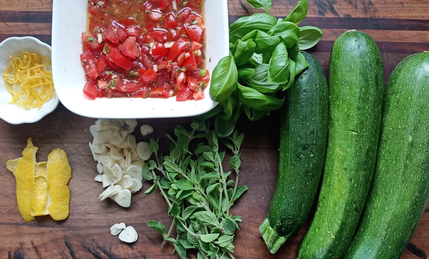 Auf einem Brett eine Schale mit der Tomaten-Salsa, Zitronenschale, viel Knoblauch, Oregano-Zweige, Basilikum und drei Zucchini