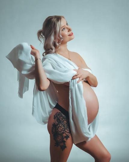 The image features a pregnant woman draped in a white cloth. She has a tattoo on her thigh and is posing in a serene manner against a neutral background.