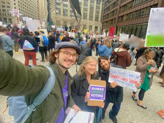 Three FairVote Illinois volunteers at a large protest in Daley Plaza, downtown Chicago holding signs that say "Ranked Choice Voting" and "Fixing Politics Starts with Ranked Choice Voting"