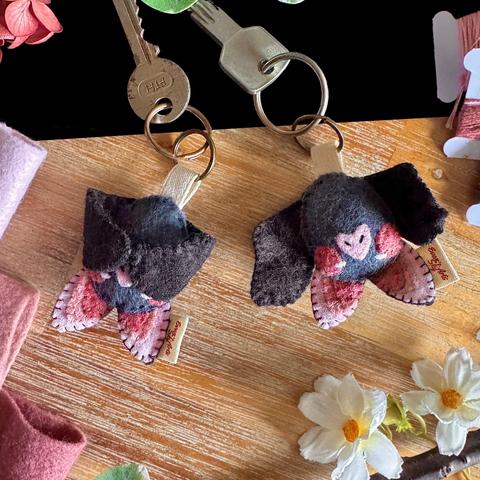 Two cute little handmade bat keychains photographed on a black and wooden background with decorative flowers and felt around them. 
The bats have big, three-toned pink ears, a small heart-shaped nose, and a dark pink blush. Their eyes are small, white and squinty. One of the bat has closed wings, held together by hidden magnets.
