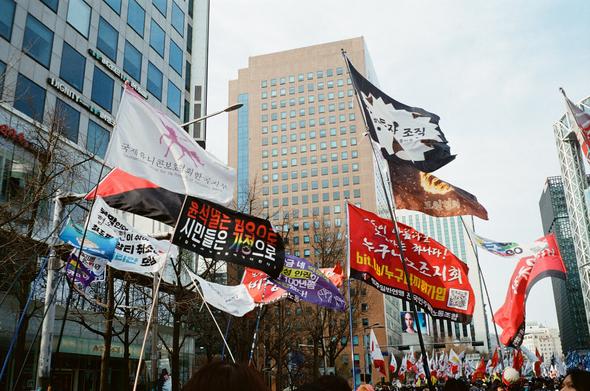 행진을 준비하는 사람들. “국제유니콘보호협회한국지부”, “윤석열은 감옥으로 시민들은 가정으로“, “누구나노조지회”, “동물권행동 카라노조” 등 다양한 깃발들이 마천루 아래 휘날리고 있다.