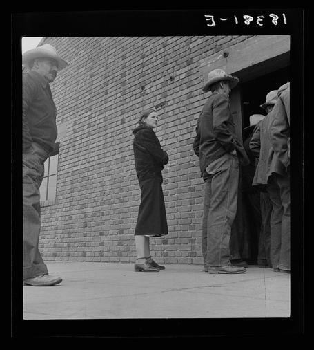 The image is a black and white photograph depicting several individuals standing in line against a brick wall, likely waiting to receive aid or assistance. The photo captures the expressions of anticipation and patience on their faces as they wait. One person stands out from this group; she appears more reserved compared to those around her.

In terms of context, Dorothea Lange was an American photographer known for documenting human rights violations during the Great Depression in America. This photograph is part of a collection that highlights the conditions faced by farm labor families at that time period, specifically focusing on their struggle with poverty and lack of resources as they sought assistance from government programs like those administered by the Farm Security Administration.

The specific individuals captured here were likely among many who relied on these services to survive during one of America's most challenging economic periods.