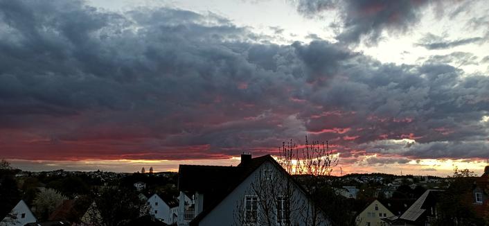 Eine dunkle Gewitterwolkenwand steht über einer Kleinstadt. Im Vordergrund mittig ein Hausgiebel, rechts davon noch die untergehende Sonne, die die Wolkenwand von unten her ein bisschen orangerot anleuchtet. Zwischen Horizont und Wolken ein schmaler Streifen Abendlicht. Vor dem Giebel ragt ein Baum seine immer noch kahlen Äste in den Himmel.