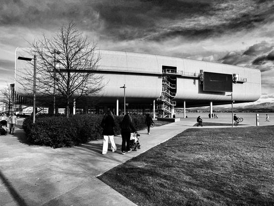 Una fotografía en blanco y negro muestra un edificio moderno y alargado con una forma curvada, similar a un barco, que se extiende horizontalmente en el encuadre. El edificio es de un solo piso y tiene una gran pantalla rectangular en su fachada.  Varias personas caminan por una plaza frente al edificio, algunas empujando cochecitos.  Un árbol sin hojas se encuentra en primer plano, a la izquierda del edificio. La escena se completa con un cielo nublado y un espacio abierto con hierba corta.