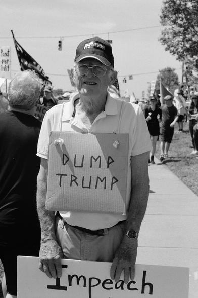 An older man with a sandwich-board-style sign saying “dump trump”