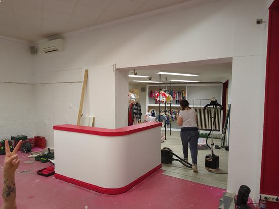 Intérieur d'un magasin de vêtements de seconde main en cours de finition avant ouverture. Les murs sont blancs et rouge. On distingue des vêtements d'enfants accrochés au fond de la boutique.