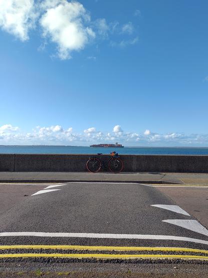 a bicycle parked against a sea wall

in the foreground a grey road with double yellow lines and a speed bump with triangles marking the 2 way lanes.

the sky is very blue with a white cloud above and a few clouds on the horizon

aligned with the speed bump and bicycle is a large cargo ship carrying a large number of containers, mostly orange

the bicycle is green with orange handlebars and orange tyres