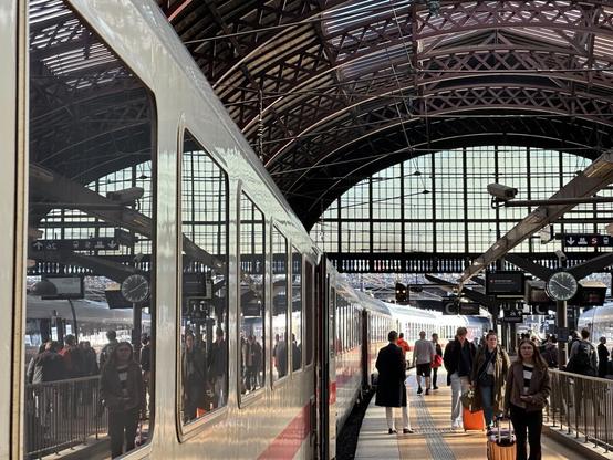 a view along a busy railway station platform. A train stands to the left, its windows reflecting the ironwork of the arched roof