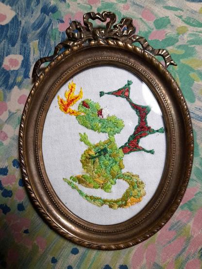 A cute green dragon with reddish-brown wings like a bat, fire-breathing. It's embroidered with needle-painting technique and framed in an oval antique bronze frame.