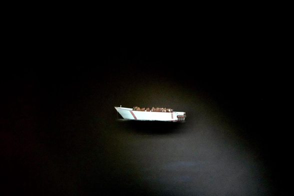 Rabenschwarze Nacht. Im Kegel eines Scheinwerfers ist ein treibendes weißes Boot von Backbord zu sehen, in dem dicht gedrängt Menschen sind. Das Boot hat drei Außenbordmotoren.