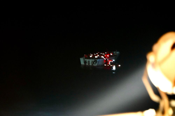Rabenschwarze Nacht. Im Kegel eines Scheinwerfers ist das treibende weiße Boot, in dem dicht gedrängt Menschen sind, von Steuerbord Achtern zu sehen. Das Boot hat drei Außenbordmotoren. Schemenhaft zu erkennen das sich bei ihm ein kleines weiteres Boot befindet. Die Menschen sind jetzt mit Rettungswesten versorgt, die teilweise orange im Scheinwerferlicht leuchten und vor allem die Reflektoren werfen das Licht deutlich sichtbar zurück. Rechts am Bildrand ragt der Scheinwerfer ins Bild, der die Szenerie ausleuchtet.