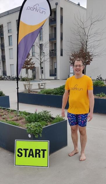 Kim-Thomas Rehmann steht vor einer Parkrunfahne. Links von ihm steht ein gelbes Schild mit dem Aufdruck Start. Kim trägt ein orangefarbiges Shirt und eine blaue Hose mit bunten Fußabdrücken. Er trägt keine Schuhe.