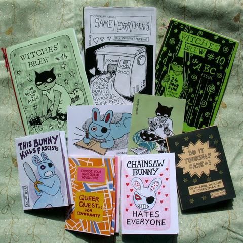 Several zines, mini-zines, and stickers spread over a green piece of fabric. Covers and images include bunnies, cats, and a sewing machine. Zine titles are Same Heartbeats, Witches' Brew, Queer Quest, Do it Yourself Care, This Bunny Kills Fascists, and Chainsaw Bunny Hates Everyone.