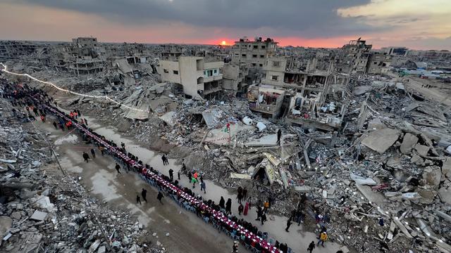 People sit for the first Iftar (breaking of the fast) of Ramadan in Rafah, southern Gaza, on 1 March, at sunset, at a long table the length of the street, surrounded by ruins & rubble.