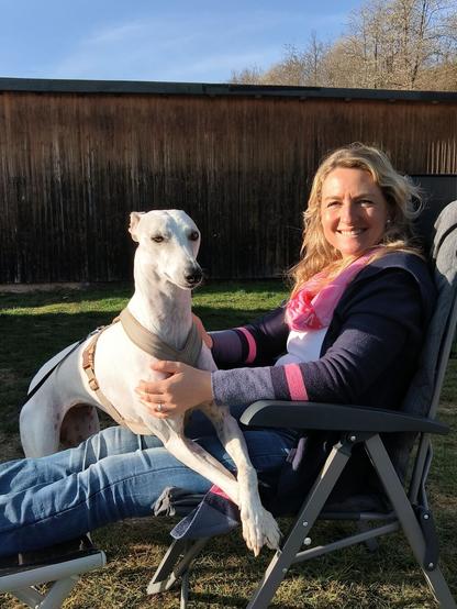 Kate sitzt auf einem Campingstuhl. Auf ihrem Schoß ist mir den Vorderbeinen Greyhound Dame Solana