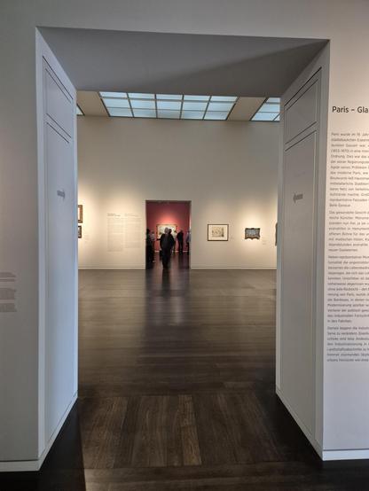 Durchblick durch einen hellen Ausstellunsraum, im Hintergrund ein rot gestrichener Raum mit vielen Besuchern.
