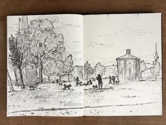 A meadow with various shadows of people and dogs on it, some benches, lots of treesbehind the meadow, an old tower with graffiti on the left side and two smaller buildings in the middle and the rigth of the picture. Buildings and the Fernsehturm in the Background.