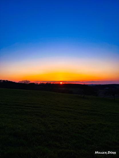 Ein ruhiger Sonnenuntergang über einer sanft geschwungenen Wiese. Die untergehende Sonne taucht den Himmel in warme Orange-, Rot- und Violetttöne. Dunkle Bäume stehen als Silhouetten am Horizont, während der Vordergrund im Schatten liegt. Die Szene wirkt friedlich, hoffnungsvoll und still.
