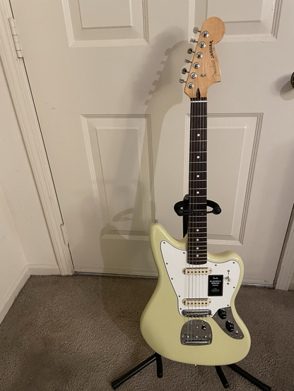 A full view of Hope, the Hialeah yellow Fender Jaguar electric offset guitar. Hope has a maple neck and headstock and a rosewood fretboard. The pick guard is white and the Fender sticker is on it. There are two single coil pickups with shark teeth style guards which reduce hum. The fretboard has pearloid dot inlays. Hope is on an onstage classic stand on the carpet in front of an off white door. Have a lovely evening!