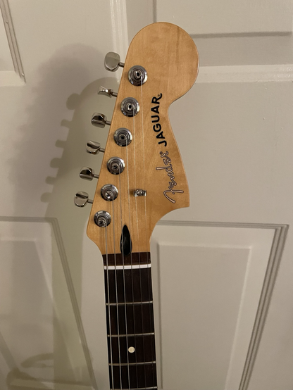 A close up of Hope’s headstock. The tuners are ClassicGear, silver colored and all on one side of the headstock. The nut is white and the fretboard joint is glossed along through the headstock. The headstock is a soft golden color with swirling wood grain. Warm and catches the light. The fender logo is clear and Jaguar is black in all caps.