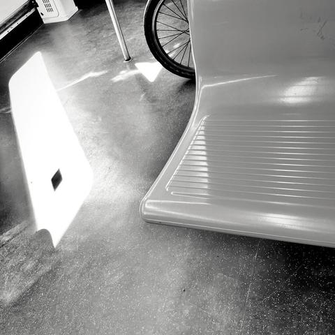 A close-up of a public transport seat and bicycle wheel, with sunlight casting a shadow on the floor, all in black and white.