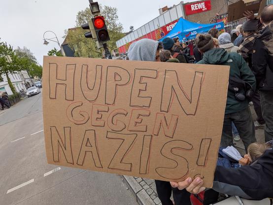 Eine Hand hält ein Pappschild mit der Aufschrift „Hupen gegen Nazis”, dahinter sind eine Straße, eine rote Ampel, ein Supermarkt und eine Menge stabile Leute zu sehen, die gegen einen fucking AfD-Stand in der Bremer Neustadt protestieren.
