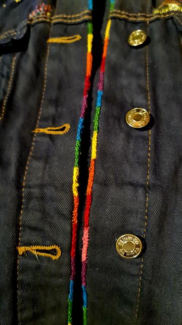 Handgearbeitetes visible mending an den Stößen der knopfleiste einer Jeansjacke. Die Flickarbeit ist beidseitig in Regenbogenfarben.