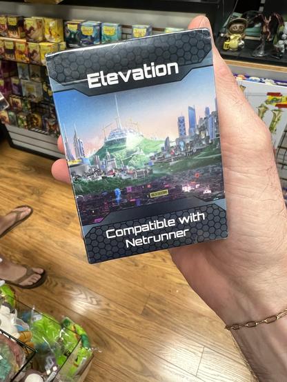 A close-up of my hand holding the box of the newest set of Netrunner cards called Elevation. In the background is the shelving and floor of a game store. The box art shows a shot of Kota Kalimantan, the fictional megacity in Borneo where the set is located. It’s got low-lying slums in the foreground, clusters of sky scrapers in the distance and behind it all, the new space elevator the city was founded to build rising into the sky with cranes around it.