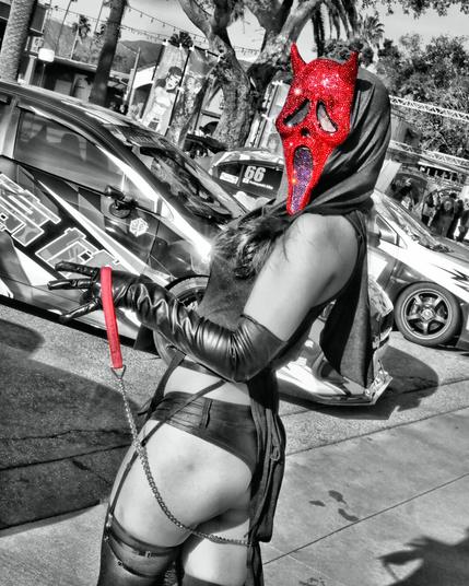 Cosplayer wearing a red ghostface mask from the movie Scream.  The mask is covered in gems which sparkle in the sunlight.  She wore a black bra under a thing dress.  Her arms are covered in long black latex gloves and used a metal chain and red collar.  Her arm is up exposing her side and armpit and the face is tilted toward the right toward the armpit.  In the background there are various automobiles with anime characters painted on them.  The image is taken in natural daylight at Anime Impulse in Pomona California on February 15, 2025.