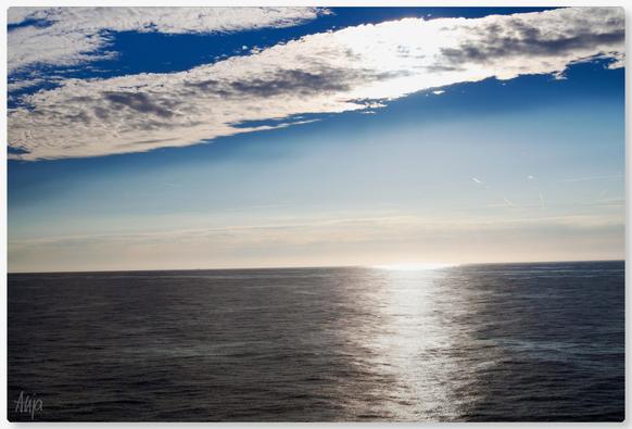 Blick von einer Fähre auf den Sonnenuntergang in der Nordsee. Ein grau-weißes Wolkenband am oberen Bildrand, darunter blauer Himmel, die Sonne versinkt am Horizont, der Lichtstreifen auf dem Wasser glänzt silbrig, alles schimmert. 

View of the sunset in the North Sea from a ferry. A grey-white band of clouds at the top of the picture, blue sky below, the sun sinking into the horizon, the strip of light on the water glistening silvery, everything shimmering.