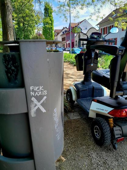 The scoot-chair is parked next to a trash can emblazoned with FUCK NAZIS, and the Shitter logo, which is a sans-serif swastika.