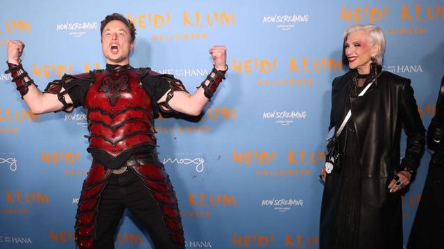 A man [Elon Musk] in a red and black armored costume poses triumphantly with his arms raised, while a woman [Musk's mother] in a stylish black outfit and short platinum blonde hair smiles beside him. The background features a blue wall with "Heidi Klum Halloween" promotional text.