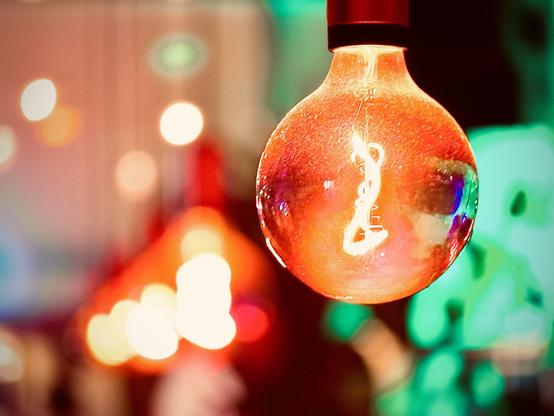 A red-orange incandescent light bulb hangs from a ceiling in a colorfully lit nighttime space, with four additional globe bulbs alight down a line in the blurry distance.