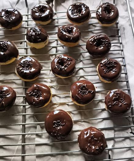 Mini Dounuts mit Schokoglasur und Schokostreusel auf einem Gitter stehend. Unter dem Gitter liegt graues Amazonpapier um den Tisch vor Tropfen zu schützen.