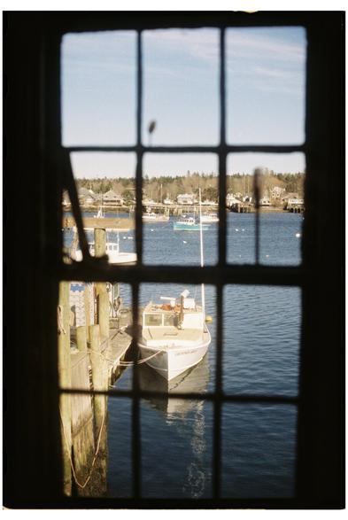 A 35mm film photo taken through a closed window. The window muntins are out of focus in the foreground and the subject of the photo, a lobster boat tied up at a float, is in focus in the background.