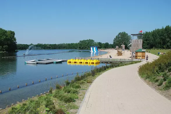 The image depicts a scenic lakeside area. There is a pathway along the water, with several yellow paddle boats lined up at a dock. In the background, there's a sandy beach area, along with some inflatable slides or structures. The setting is surrounded by greenery, with trees visible, and the sky appears clear and blue. It looks like a recreational place suitable for swimming and outdoor activities.

Image Credits: Wikimedia / Paul Hermans / CC BY-SA 3.0