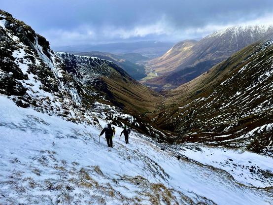 Two walkers with axes and helmets walk up from Glencoe, partially snow covered hills in the background with a combination of sun and clouds dappling the mountains.