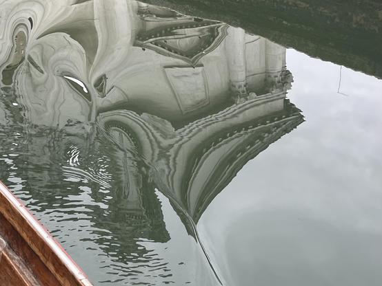 Colour photo, taken aboard a boat, looking down into silvery green canal water. The image is framed in the upper right corner by the reflection of a low brick canal wall, and in the lower left corner by the edge of the boat, made of bright brown shiny wood. Reflected between them is a monumental marble building, with columns and crenellations, perfectly mirrored in the water which is completely still and flat, apart from along the boat's edge in the lower left corner where it is rippled and distorted by the boat's gentle wake. The overall effect of the image is one of a sort of optical illusion, reality being invaded and slightly distorted by a tiny vortex from elsewhere, and for me feels like it's asking us a question about what we perceive and how we perceive it.