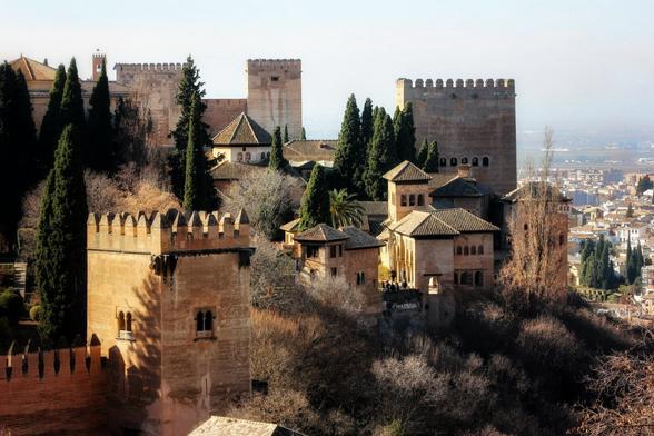 A high-angle shot shows the ancient, reddish-brown stone walls and towers of the Alhambra palace complex nestled on a hill amongst trees, with a hazy cityscape visible in the background.