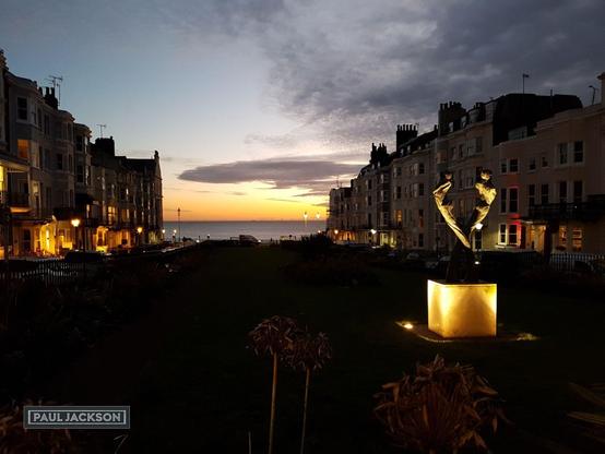 Twilight descends upon New Steine Gardens in Brighton, England.

The sky is a beautiful blend of colors, with the last light of the sun painting the horizon over the sea in warm oranges and yellows. Above that, the sky transitions into softer blues and grays, with some interesting cloud formations adding texture.

The view looks down towards the sea front with a row of tall four or five storey houses on either side of the picture. A narrow well kept garden, now in darkness, leads down the centre of the picture. Flanking the gardens on either side are the elegant, tall buildings typical of Brighton's architecture. Many of their windows are lit up, casting a warm glow onto the street and the gardens below. The streetlights also contribute to this gentle illumination.

In the foreground, the gardens themselves are barely visible, with some plants and flowerbeds. A prominent feature is the illuminated sculpture on a pedestal, drawing the eye. It adds a touch of artistic flair to the scene.

A peaceful and serene scene, capturing a quiet moment in this vibrant seaside city.