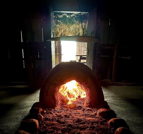 This image shows a traditional wood-fired oven, often used for baking bread or pizza. The fire inside the oven is burning brightly, illuminating the dome-shaped interior. The construction appears to be made of a clay or brick material, typical for this type of oven, which retains high heat for efficient cooking. The background shows a rustic setting with a faint view of some plants hanging and a doorway leading outside. The scene is warmly lit, suggesting a cozy, rural or traditional kitchen environment.