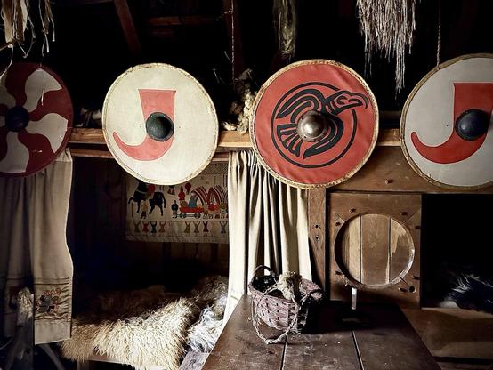 The image depicts an interior scene reminiscent of a traditional or historical setting, possibly themed around a Viking or medieval culture. Featured prominently are several round shields with various designs, including symbols and patterns typical of a Norse or Old Germanic style. These shields are mounted on a wall adorned with curtains and other rustic decorations. Below the shields, there appears to be a wooden bench covered with a fur throw, next to which sits a small woven basket. The overall ambiance suggests a display or a reconstruction found possibly in a museum or a cultural heritage site. This setting serves both as an educational exhibit and an atmospheric representation of historical life.