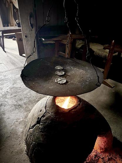 This image depicts a traditional cooking setup, likely inside a historical or rustic setting. There's an iron cooking platter suspended by chains over an open hearth, capturing the glow of burning firewood beneath. The platter has three small buns or dough pieces on it, being heated or cooked by the heat from the fire below. The room has a dimly lit, cozy atmosphere with wooden furniture and a rustic vibe, possibly suggesting a setting in a historical cottage or a recreation of an old-fashioned kitchen.