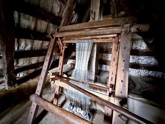 The image shows a traditional wooden weaving loom inside a rustic building, likely an old cabin or shed. The loom is equipped with a warp of threads through which the weave is being formed, creating a fabric with a simple striped pattern. The surroundings with wooden logs and dim lighting add to the historical or antiquated feel of the scene. This loom is typical of those used historically for making textiles by hand.