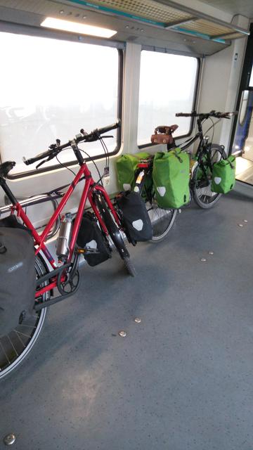 Ein rotes Patria Fahrrad namens Leo mit dunkelgrauen Gepäcktaschen vorne und hinten sowie ein Englisch-grünes Wanderer Fahrrad namens Emmi, mit hellgrünen Gepäcktaschen vorne und hinten sind im Fahrradabteil eines Zuges festgezurrt.